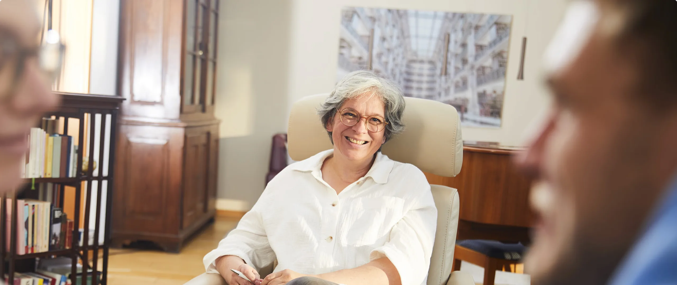 Dorothee Bremer smiling in an armchair in her practice, a bookshelf and a glass-fronted cabinet behind her, seen past the shoulders of two people sitting opposite her